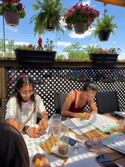 Two women practicing henna at a patio table under hanging ferns and flowers