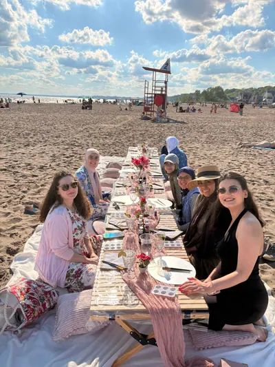 Friends seated around a long picnic table on the beach, palettes and glasses set for a paint and sip